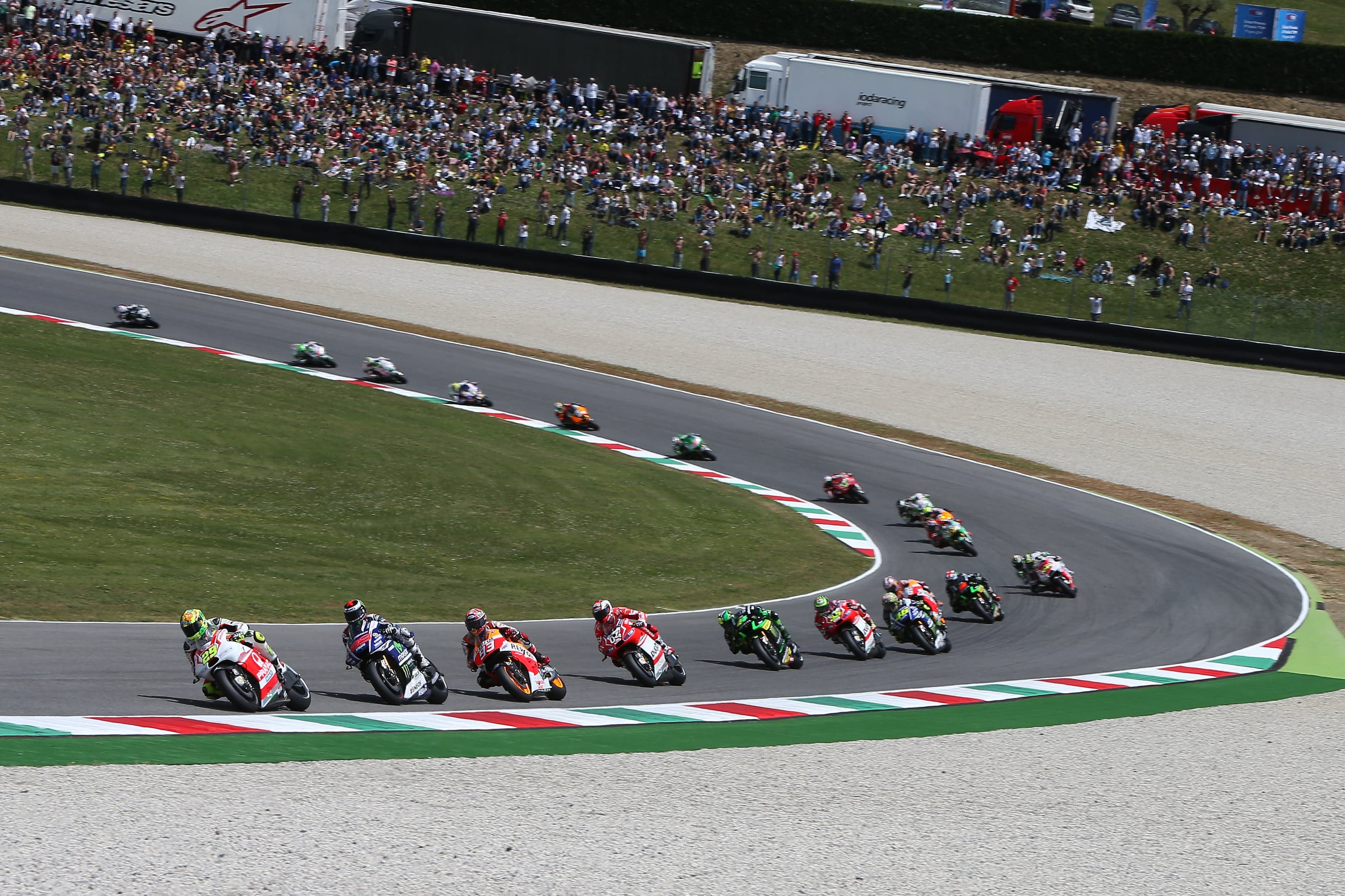 Peloton de MotoGP étiré dans une grande courbe aux vibreurs tricolores du circuit du Mugello, talus de spectateurs en arrière-plan
